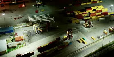 A container terminal at night, seen from above: stacks of yellow and red containers under floodlights, straddle carriers between them, and a road and a rail line running along the edge of the yard.