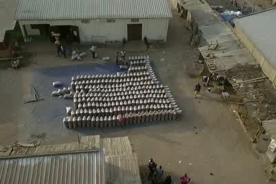 Seen from above: several hundred filled sacks stood upright in rows on the ground of a walled yard, one person walking between them, corrugated roofs along the near edge.